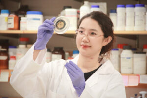 A female scientist examines a lab specimen. She is wearing a white lab coat and protective gloves.