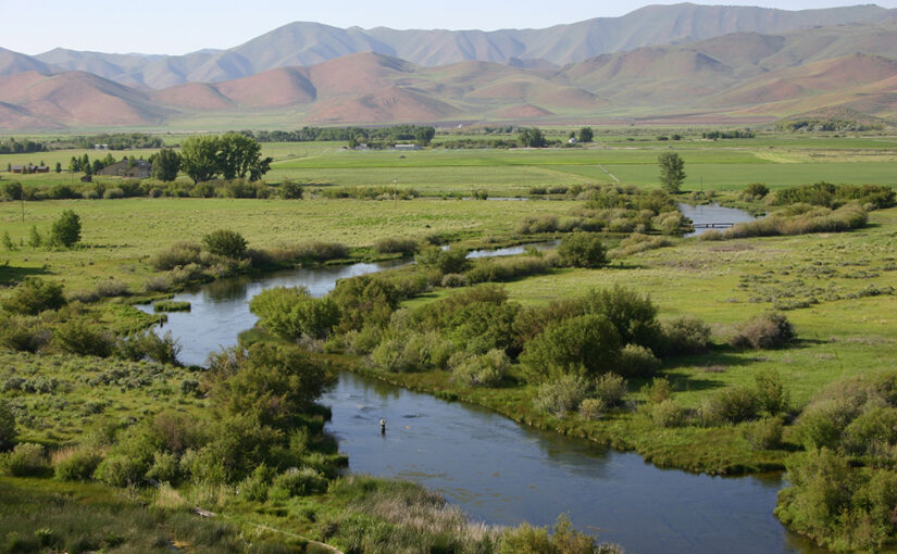 A stream meanders through green cropland with mountains in the distance