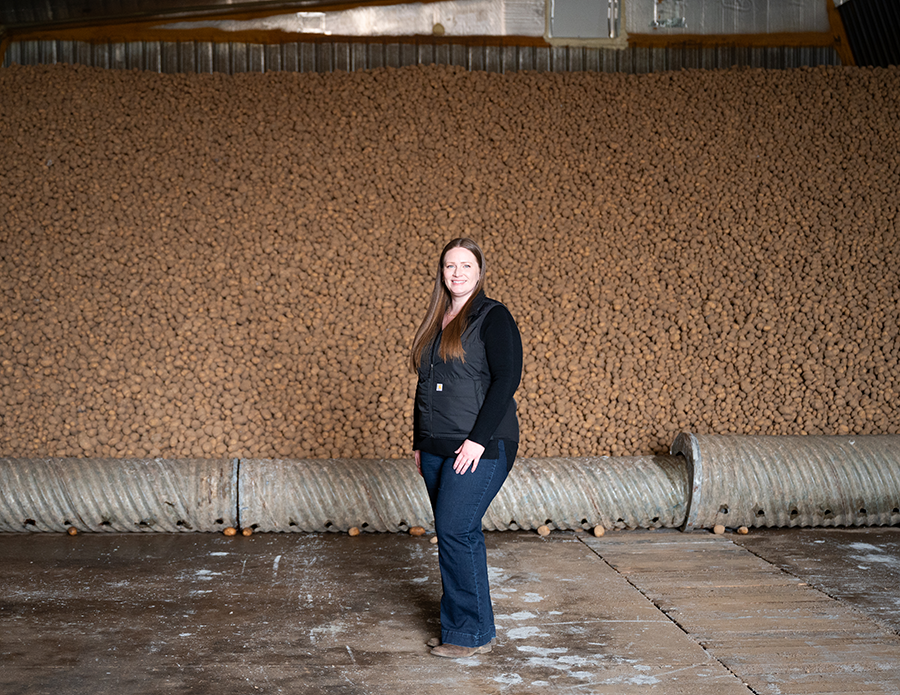 A woman in jeans and work boots poses in front of a pile of stored potatoes
