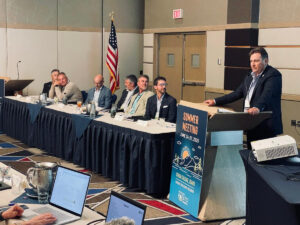 A man in a suit speaks at a podium as other men look on