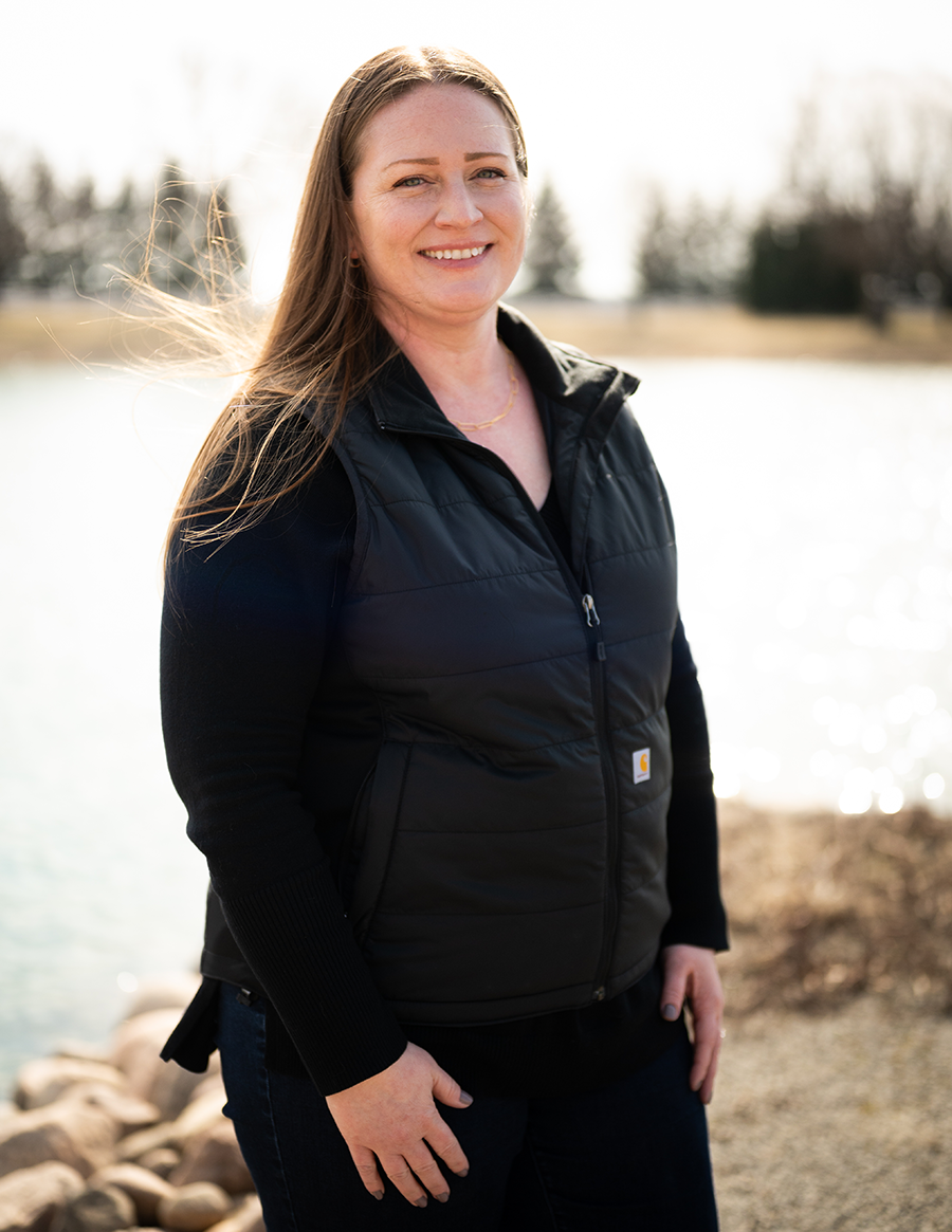 A woman stands, smiling, in front of a pond, her long brown hair lifting in a breeze
